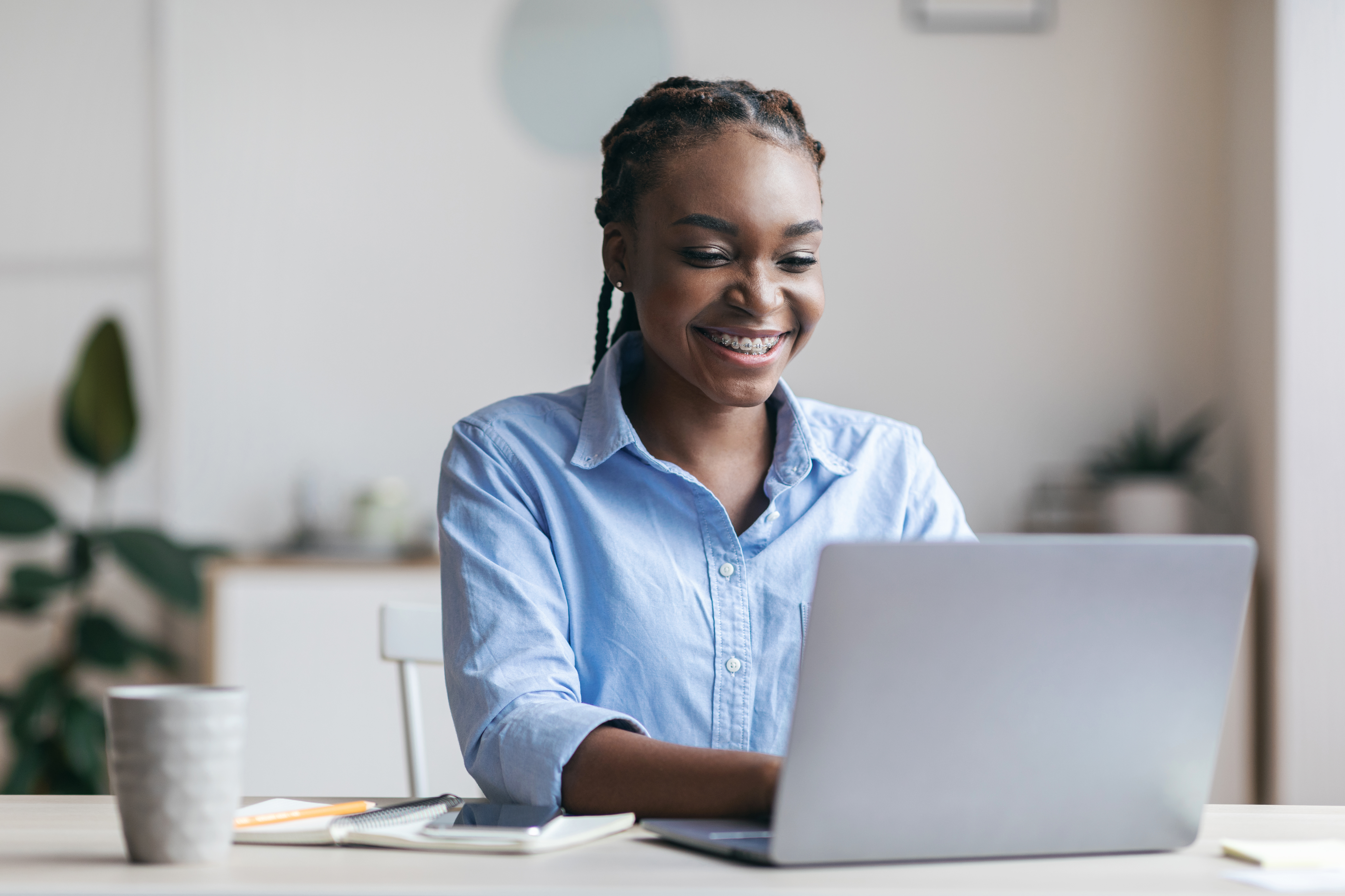 mujer morena sonriendo y trabajando en su computador portátil de color plateado y una taza con un cuaderno en su escritorio mujer morena sonriendo y trabajando en su computador portátil de color plateado y una taza con un cuaderno en su escritorio